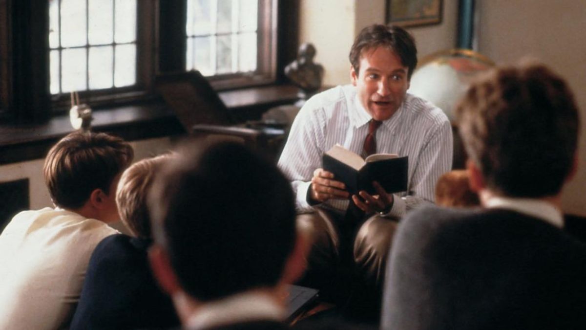 Mr. Keating (Robin Williams) sits in a classroom, holding an open book and speaking animatedly to a group of male students gathered around him.
