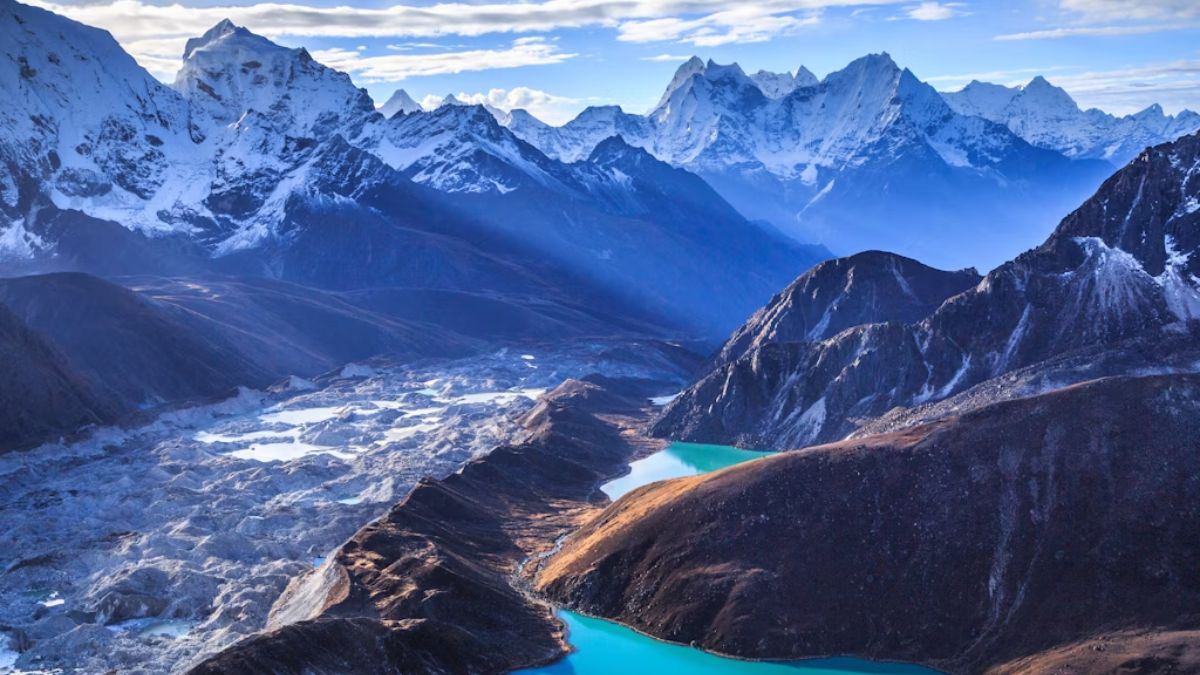 Panoramic view of the Ngozumpa Glacier and vibrant turquoise Gokyo Lakes nestled within a majestic, snow-capped Himalayan mountain range under a partly cloudy sky.