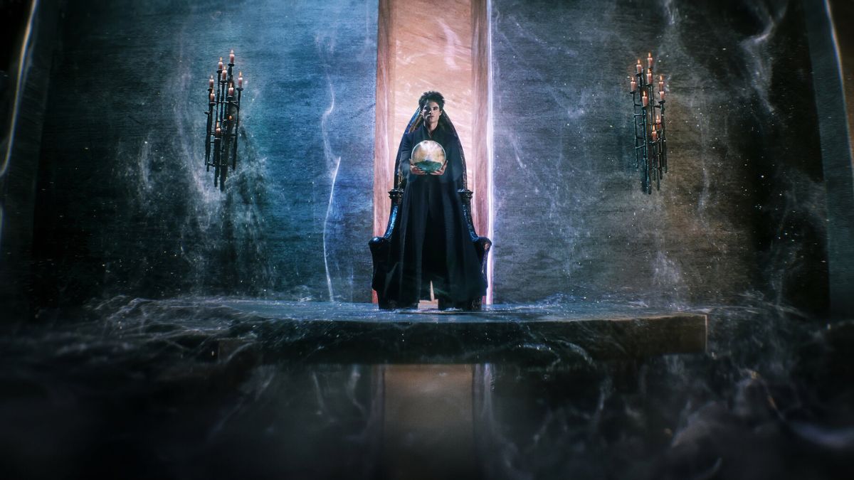 A dark, ethereal figure, identified as Dream of the Endless (Tom Sturridge), sits on a throne-like structure, holding a glowing orb. The setting is a mystical, shadowy realm with an otherworldly atmosphere, featuring tall, candelabra-like structures on either side and swirling mists at the base.