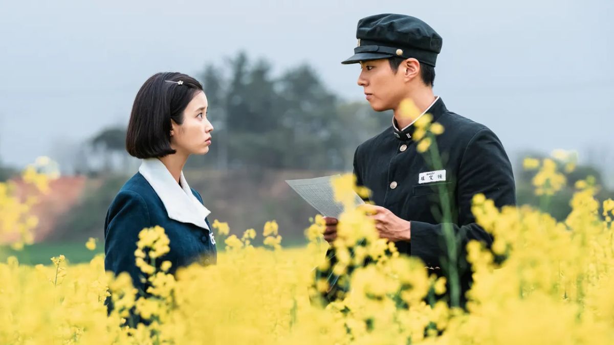 A young woman and a young man, dressed in school uniforms, stand facing each other in a vibrant field of yellow flowers. The man holds a piece of paper, and both look intently at each other.