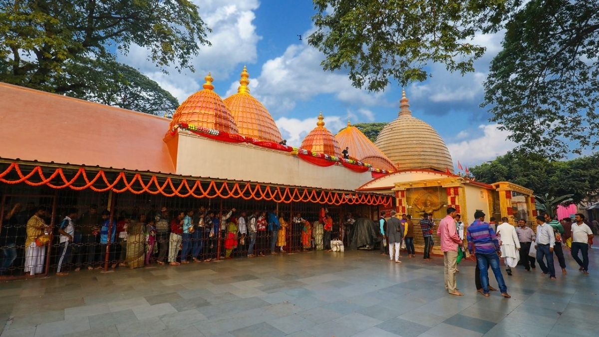 A wide exterior view of the Kamakhya Devi Temple complex in Guwahati, Assam, showing its distinctive beehive-shaped domes, a long queue of devotees, and surrounding trees under a clear blue sky.