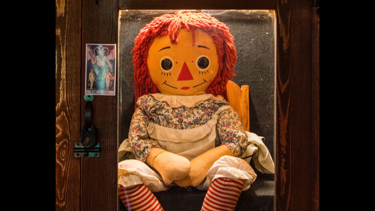 A close-up of the real Annabelle doll in its specially constructed, reinforced glass case at the Warren Occult Museum in Monroe, Connecticut, with a prominent "WARNING" sign.