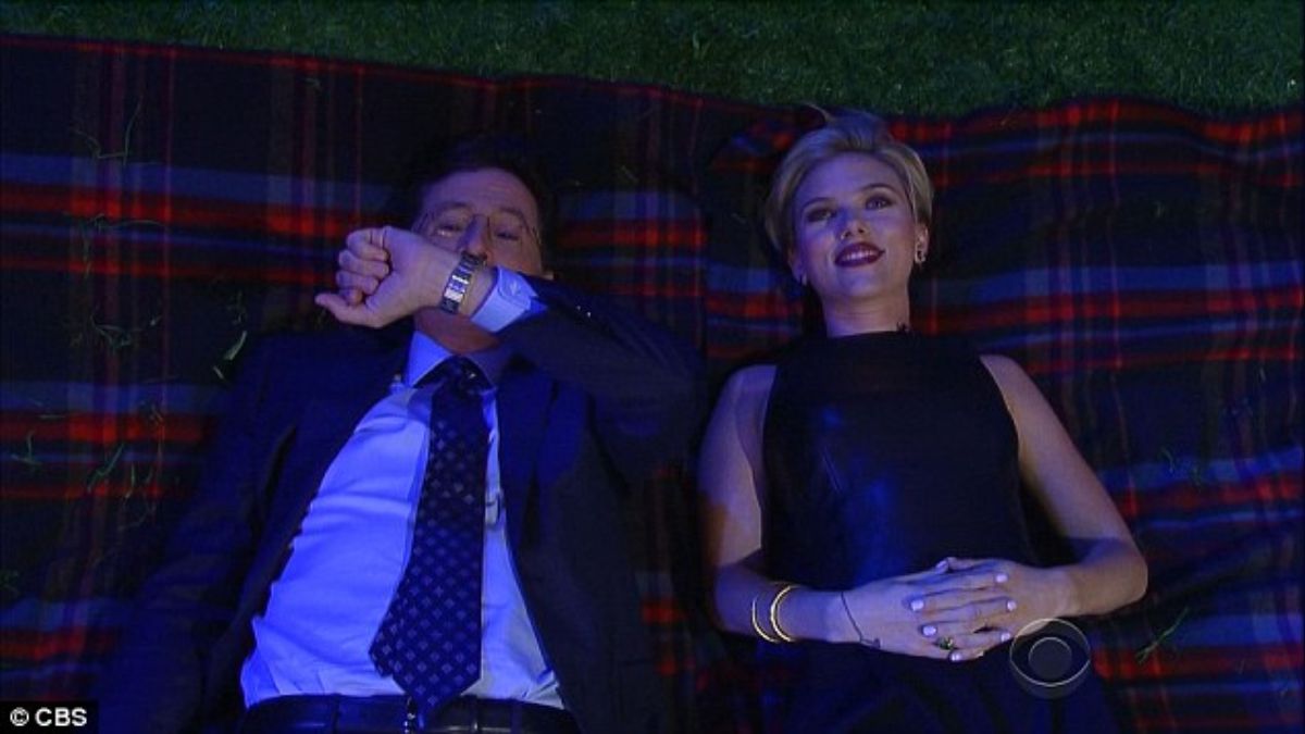 Scarlett Johansson, with short blonde hair and a black dress, smiling while lying on a plaid blanket next to Stephen Colbert, who is covering his mouth, during her appearance on "The Late Show.