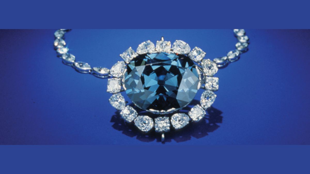 A studio photograph of the Hope Diamond, a brilliant blue diamond, set in a necklace with a surround of white diamonds. The central stone is a cushion cut, and the surrounding diamonds are a mix of different cuts. The necklace is displayed against a solid blue background.