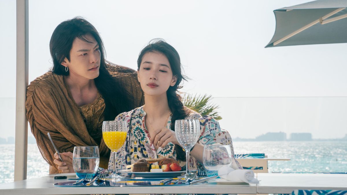 Bae Suzy and Kim Woo-bin sit at a table with food and drinks overlooking a body of water in Dubai, from the Netflix K-drama Genie, Make a Wish. Bae Suzy looks down at her hands while Kim Woo-bin looks at her with a serious expression.
