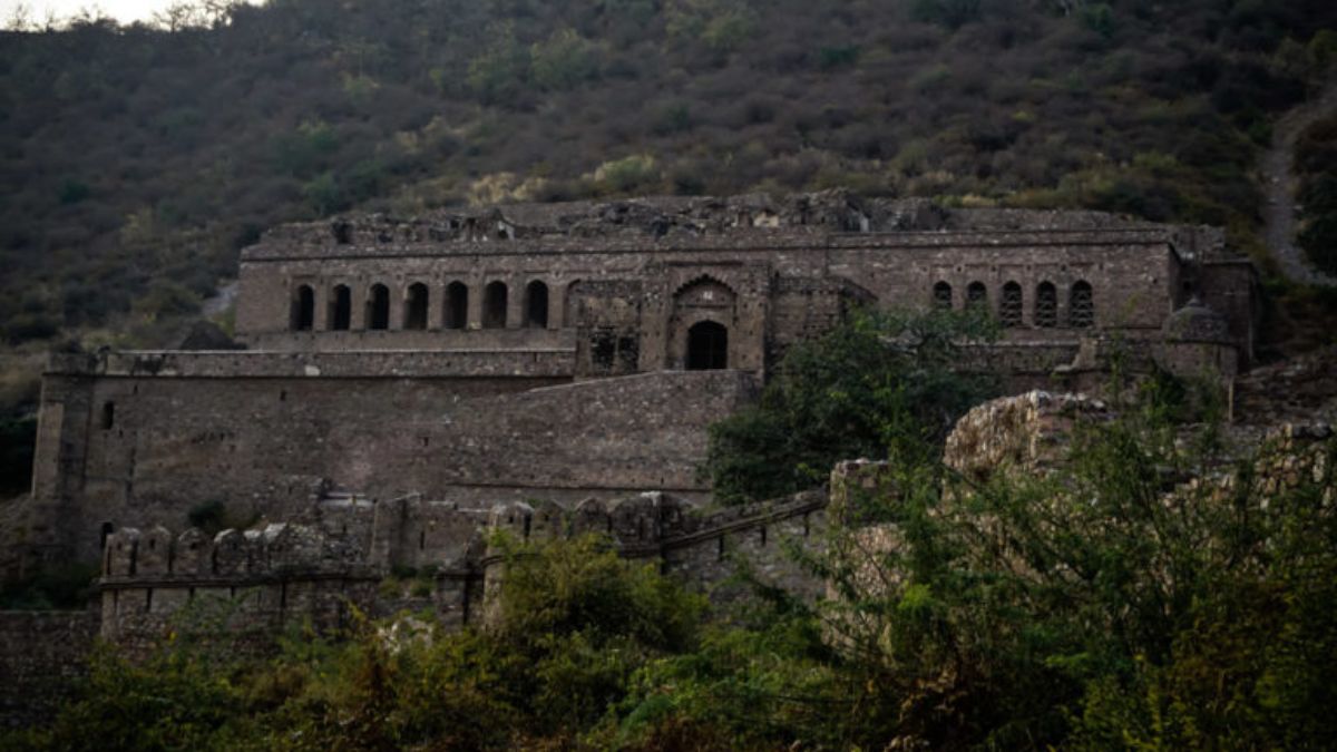 A close-up of a large, ruined stone building within Bhangarh Fort, Rajasthan, with archways and crumbling walls on a hillside.