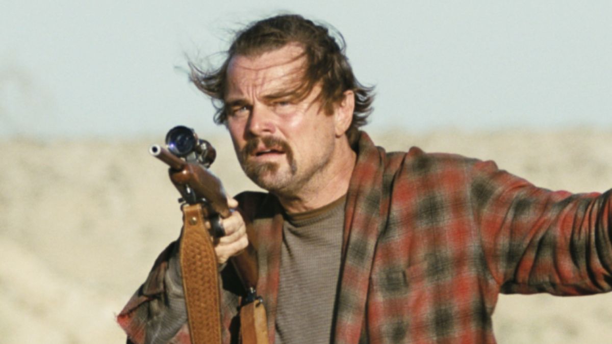 Leonardo DiCaprio, with a scruffy beard and long hair, wears a red and black plaid shirt and holds a scoped rifle, looking intently into the distance in a desert-like setting.