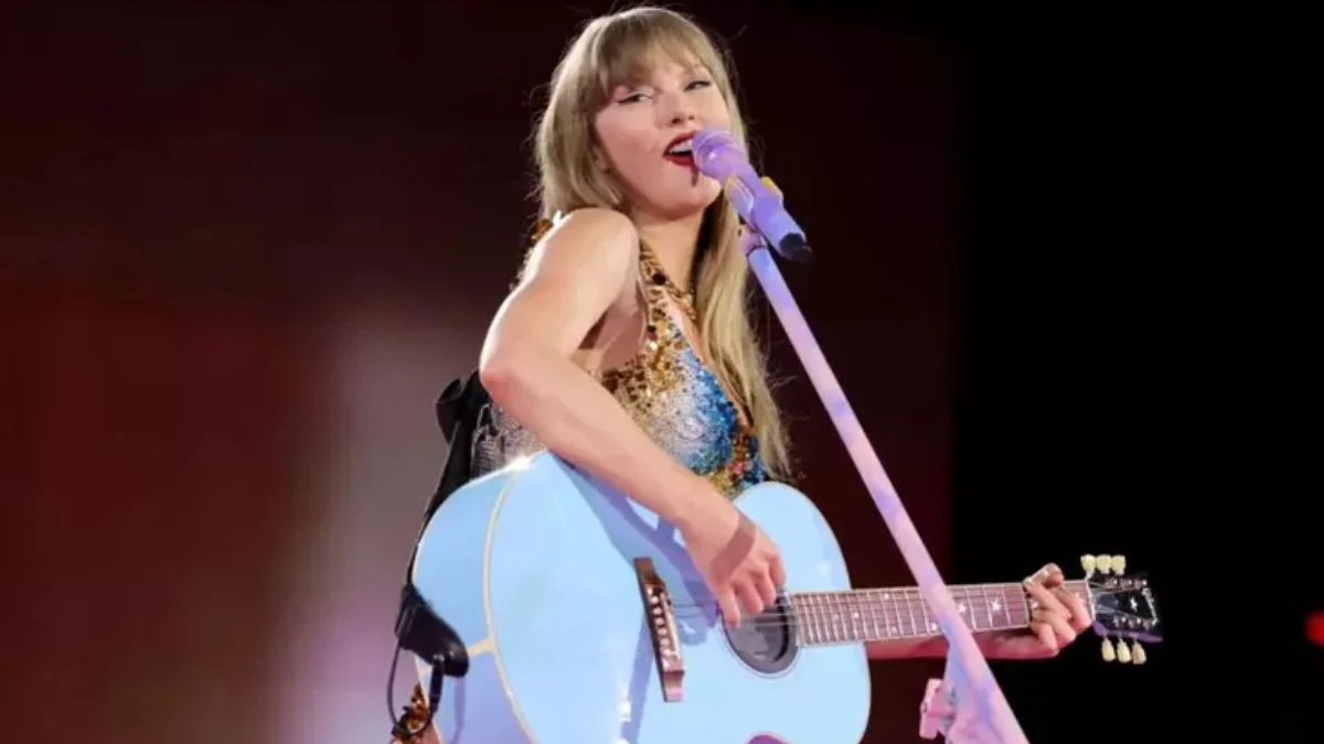 Taylor Swift performing live on stage during The Eras Tour, wearing a sequined outfit and playing a light blue acoustic guitar.