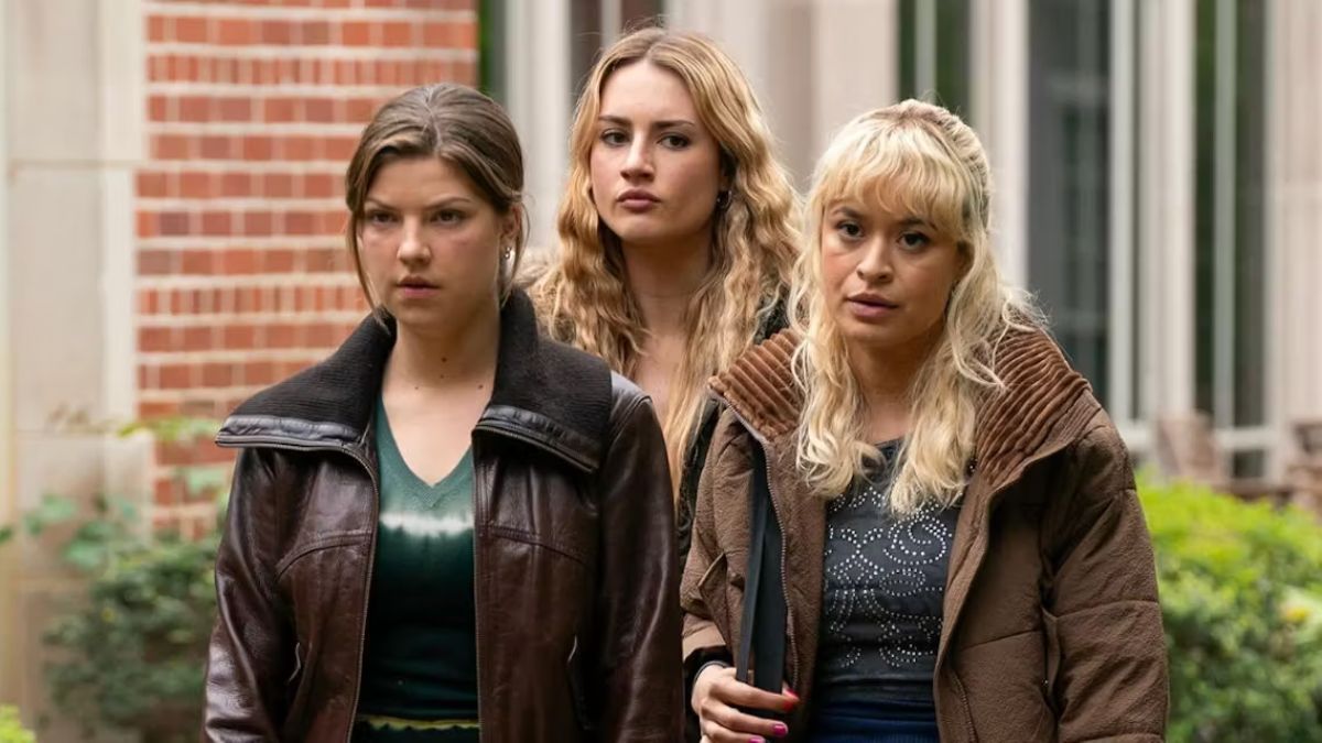 A still from Tell Me Lies featuring three young women (likely Lucy, Pippa, and Bree), two wearing leather jackets, looking serious while standing outdoors against a brick and stone building.