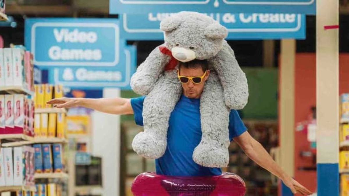 Channing Tatum as Jeffrey Manchester balancing a giant gray teddy bear on his shoulders in a toy store aisle, showcasing his secret life in the Toys "R" Us in the movie Roofman.
