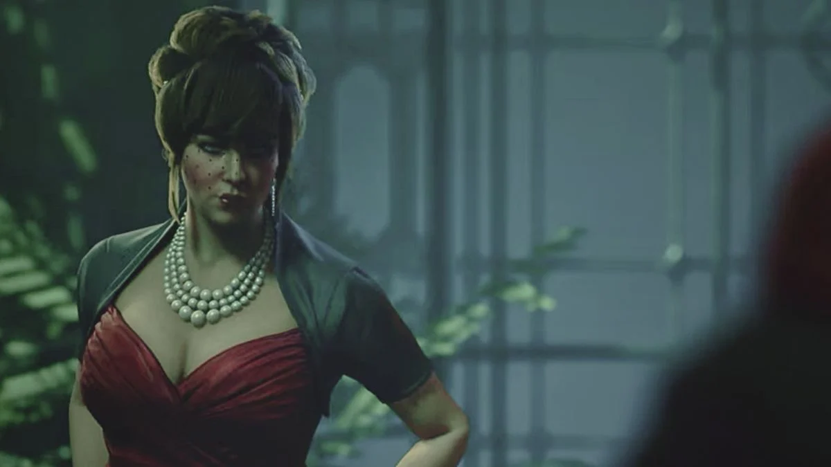 Female Kindred character in a red dress with a low neckline, a grey bolero jacket, and a striking three-strand pearl necklace, looking over her shoulder in a dimly lit, ornate interior.