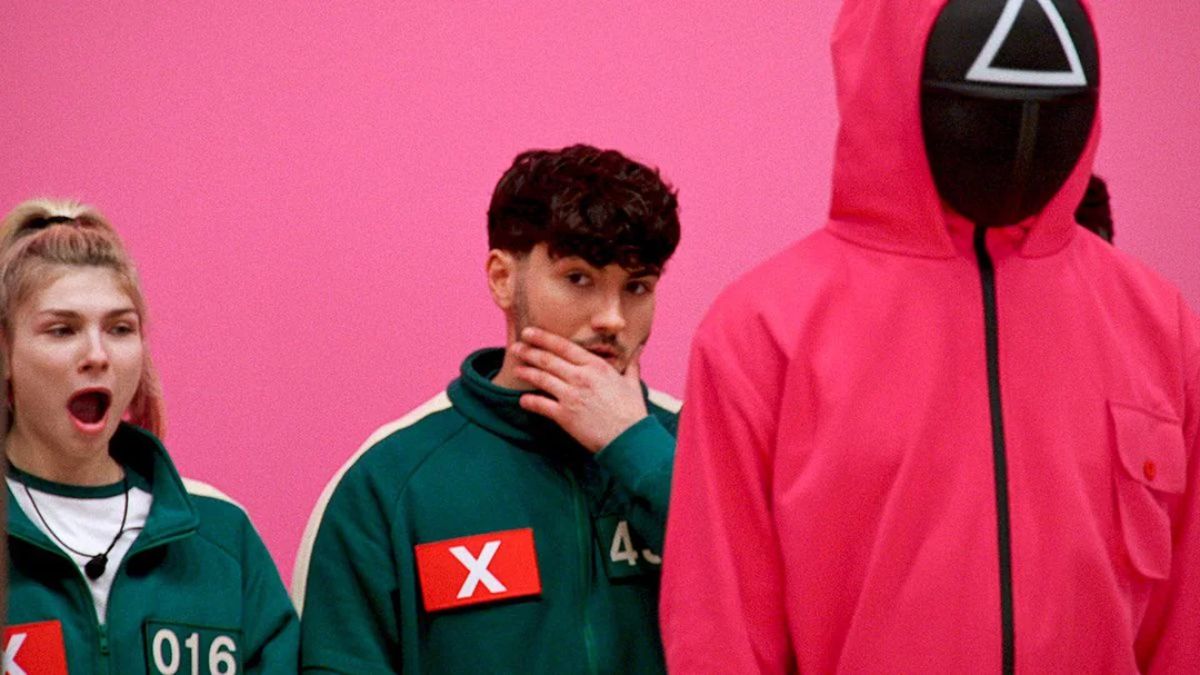 A contestant (Player 016) on the left looking shocked with her mouth open, standing next to a male contestant, while a guard in a pink jumpsuit and triangle-mask stands on the right.