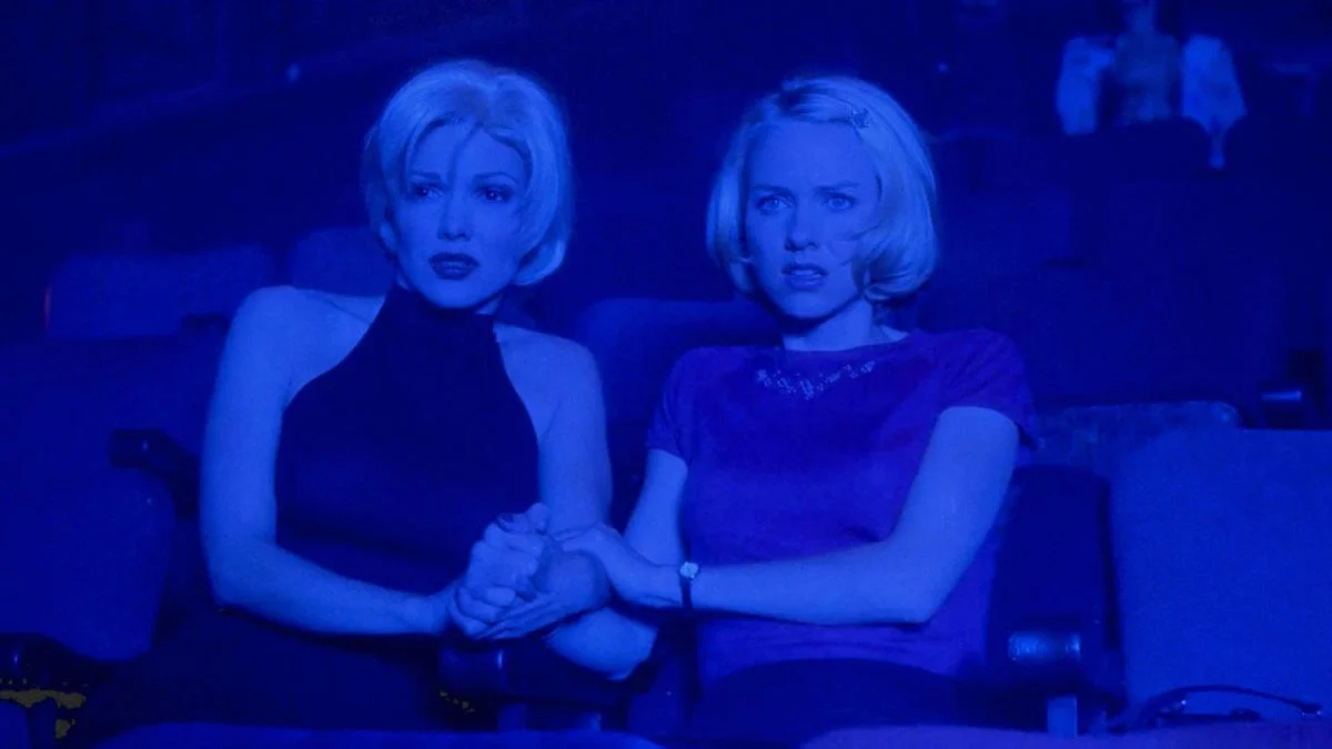 Naomi Watts and Laura Harring in a dark theater (Club Silencio) holding hands under blue light, symbolizing the collapse of illusion.