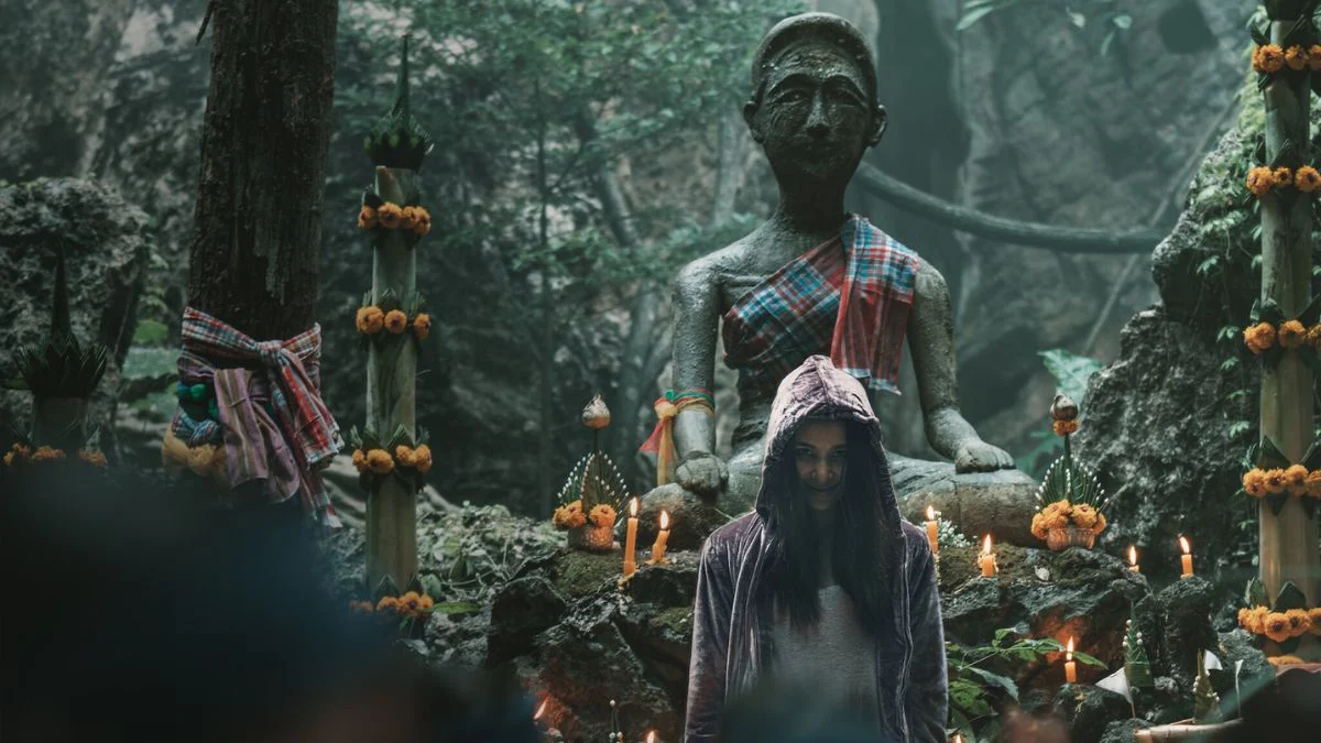 Mink, wearing a hoodie and a menacing smile, stands in a ritual space with a dark background and a stone statue behind her, hinting at her possession.