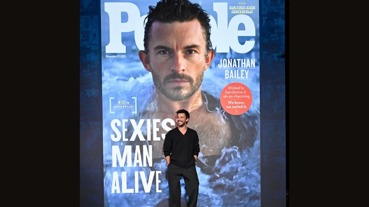 Jonathan Bailey stands smiling in front of the giant reveal of his People Magazine Sexiest Man Alive 2025 cover, which features a close-up, water-themed photo.