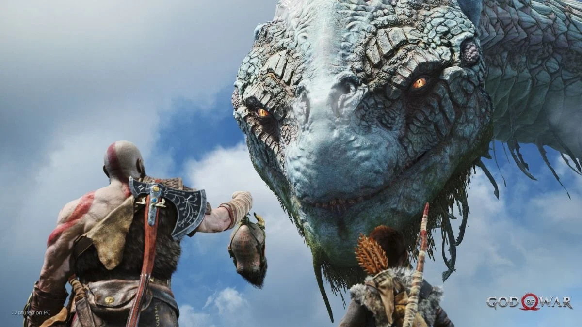 Kratos and Atreus stand before the massive head of the World Serpent, Jörmungandr, against a cloudy blue sky in God of War.