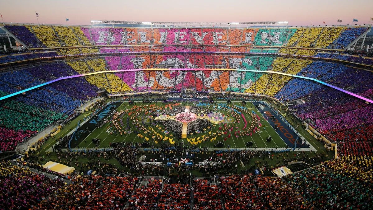 A high-angle view of a crowded NFL stadium during a Super Bowl halftime show with "BELIEVE IN LOVE" spelled out in the stands.