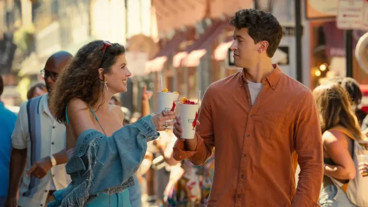 Emily Bader as Poppy and Tom Blyth as Alex walking through a sunny street with drinks in People We Meet on Vacation.