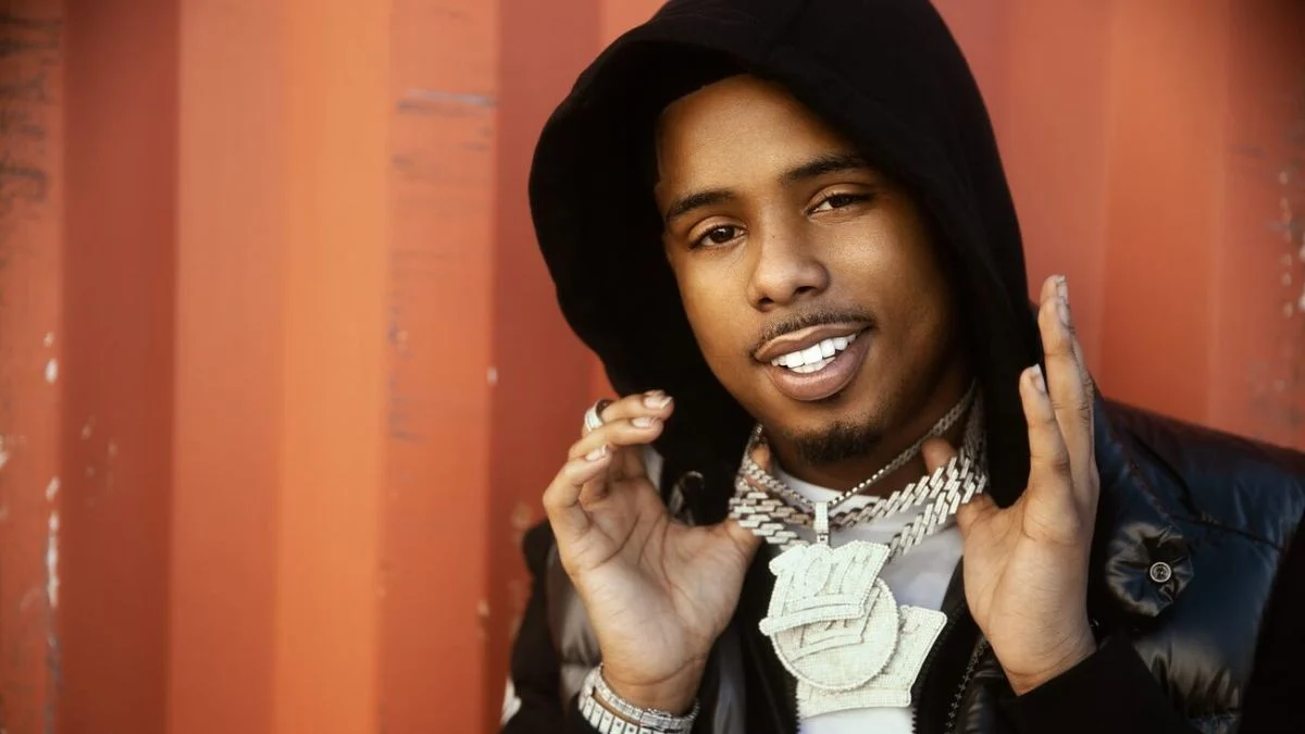 Rapper Pooh Shiesty wearing a black hoodie and a large 1017 diamond chain while smiling.