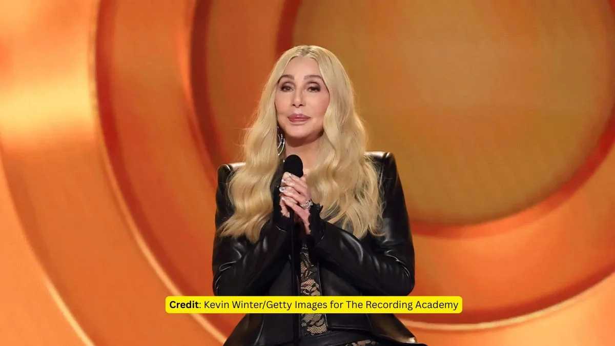 Cher standing at a microphone on the 2026 Grammy stage in a black leather jacket and lace ensemble, accepting her Lifetime Achievement Award.