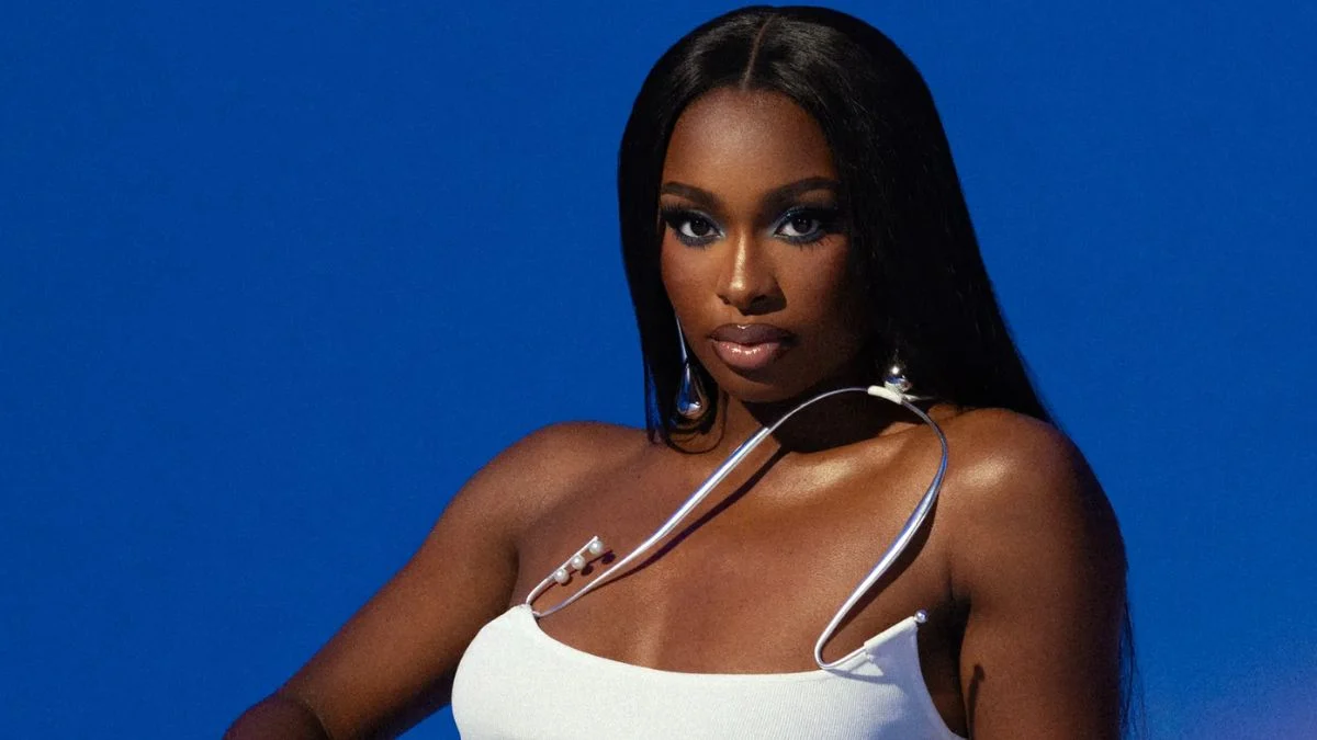 Coco Jones wearing a white halter top and silver jewelry against a deep blue background.