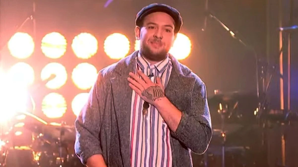 Dean Franklin, former The Voice UK contestant, performing on stage in a grey jacket and flat cap.