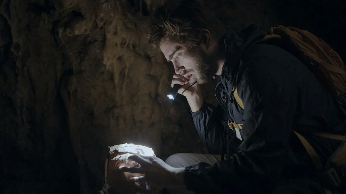 A man with a backpack using a flashlight to examine a mysterious object inside a dark cave.
