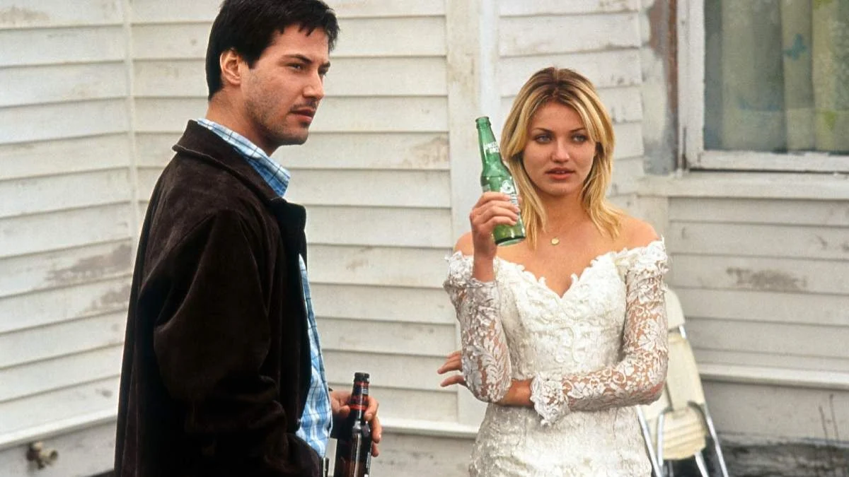 A young Keanu Reeves in a dark jacket and blue plaid shirt standing next to Cameron Diaz in a white lace off-the-shoulder dress holding a green glass bottle.