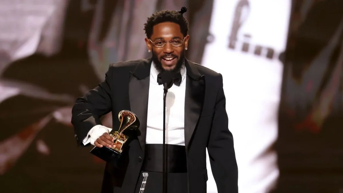 Kendrick Lamar wearing a black tuxedo and glasses, smiling while holding a Grammy Award on stage at the 68th Annual Grammy Awards in 2026.