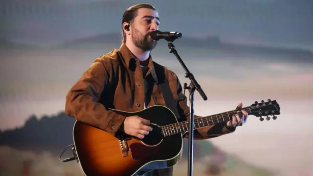 Noah Kahan performing live on stage with an acoustic guitar and a microphone during a concert.
