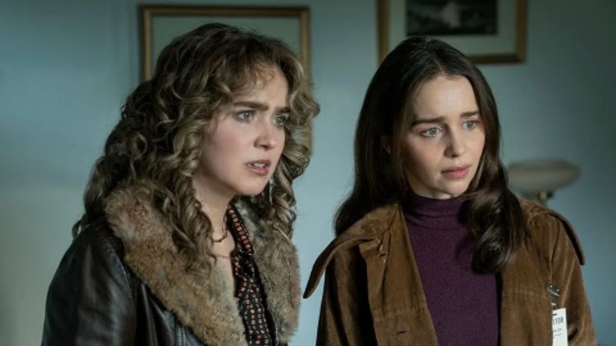Haley Lu Richardson as Twila Hasbeck and Emilia Clarke as Bea Grant looking concerned in the 1977 Moscow setting of the Peacock series Ponies.