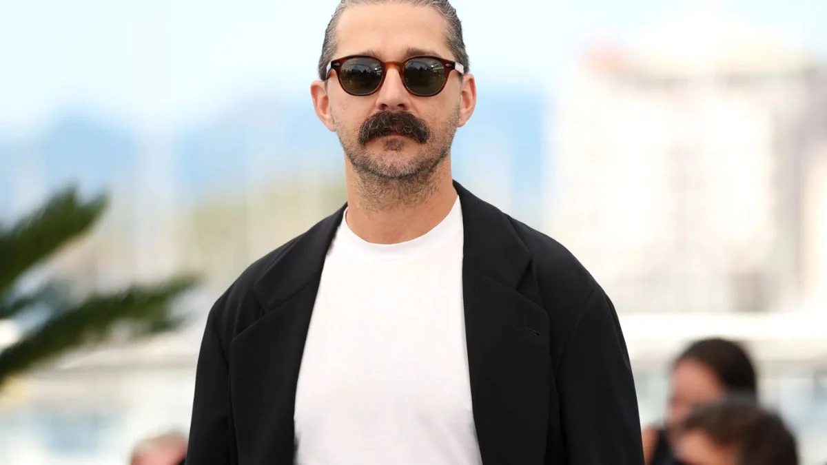 Actor Shia LaBeouf wearing sunglasses, a white t-shirt, and a black blazer with a prominent mustache.