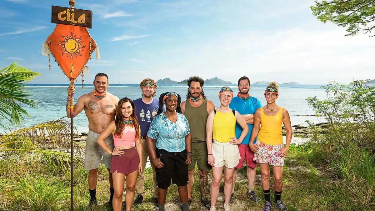 The Cila tribe from Survivor 50 standing on a beach in Fiji, featuring Cirie Fields, Ozzy Lusth, Jenna Lewis-Dougherty, and others next to their orange tribe flag.