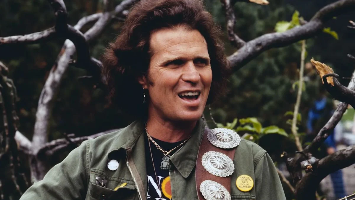 Country Joe McDonald wearing a green military-style jacket and a guitar strap with silver conchos, smiling during a performance.
