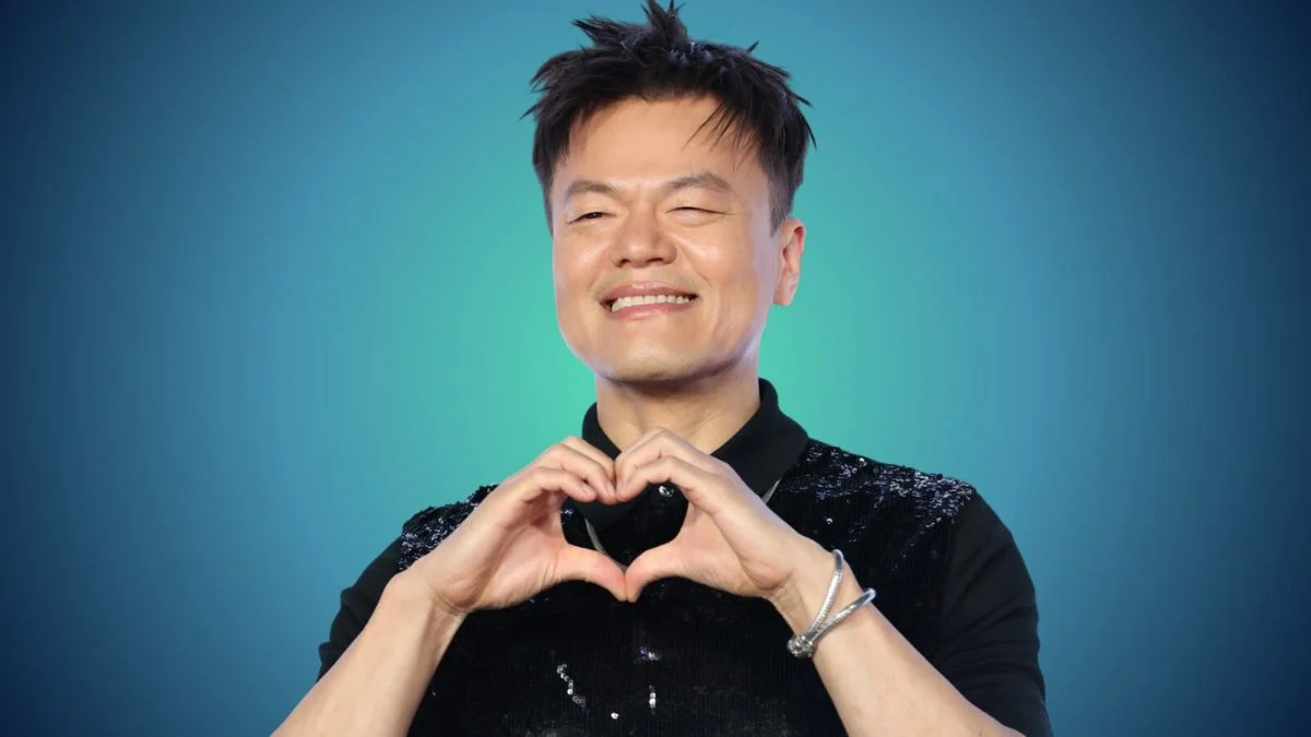 Park Jin-young (J.Y. Park) smiling and making a heart gesture with his hands against a blue gradient background.
