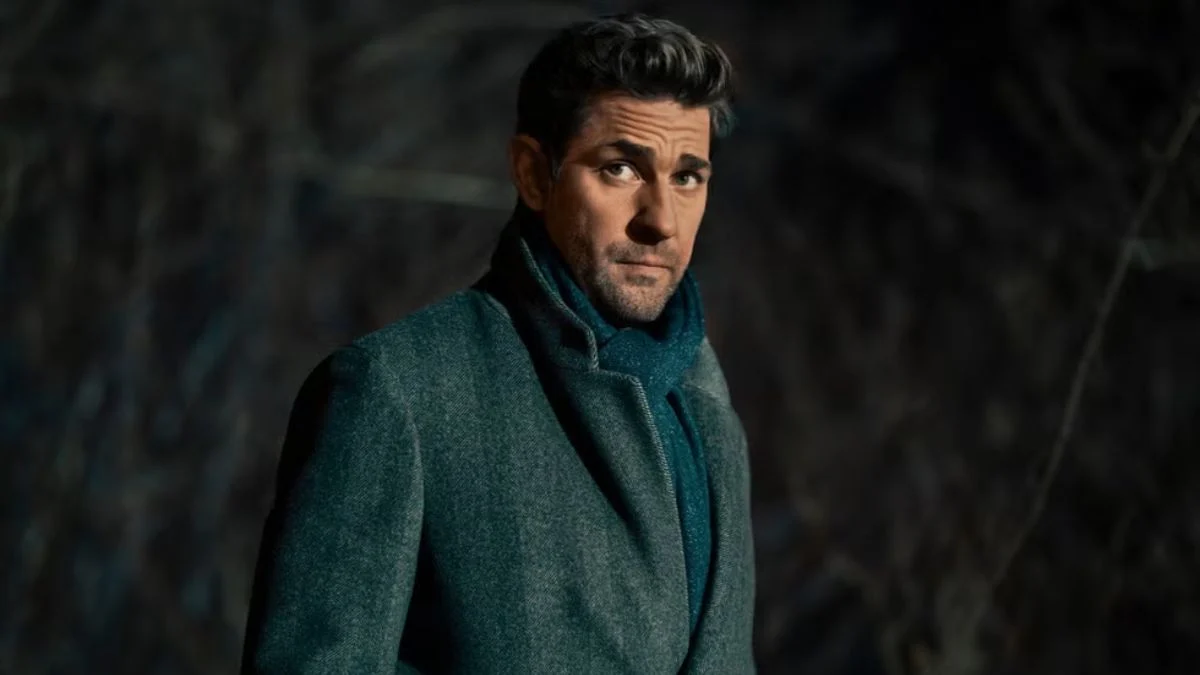 John Krasinski as Jack Ryan wearing a grey coat and blue scarf in a dark, dramatic setting.