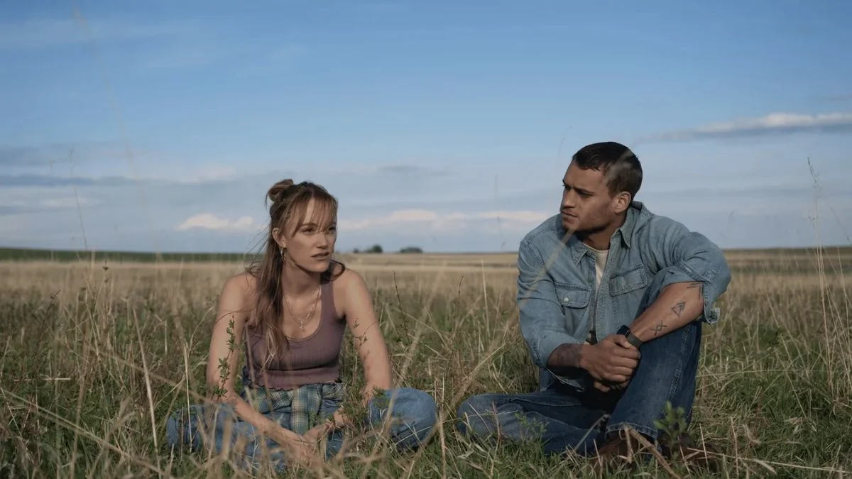 Maika Monroe as Kenna Rowan and Tyriq Withers as Ledger Ward sitting in a grassy field in a scene from the Reminders of Him movie.