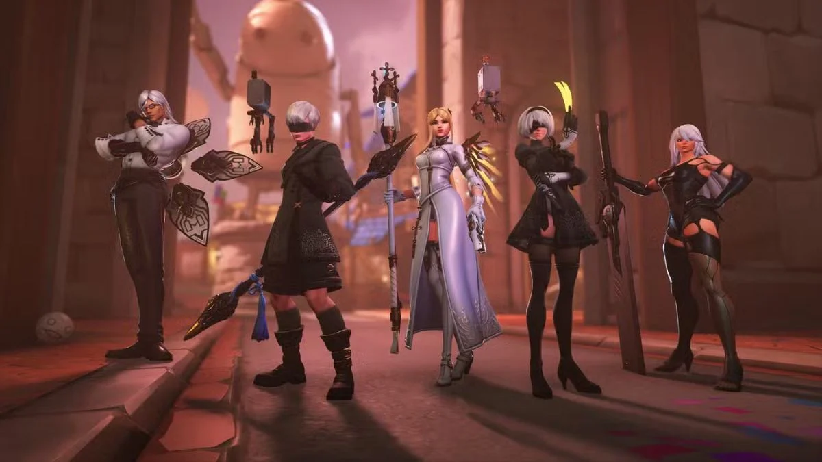 Five Overwatch heroes (Lifeweaver, Wuyang, Mercy, Kiriko, and Vendetta) wearing NieR: Automata collaboration skins standing together in an urban environment.