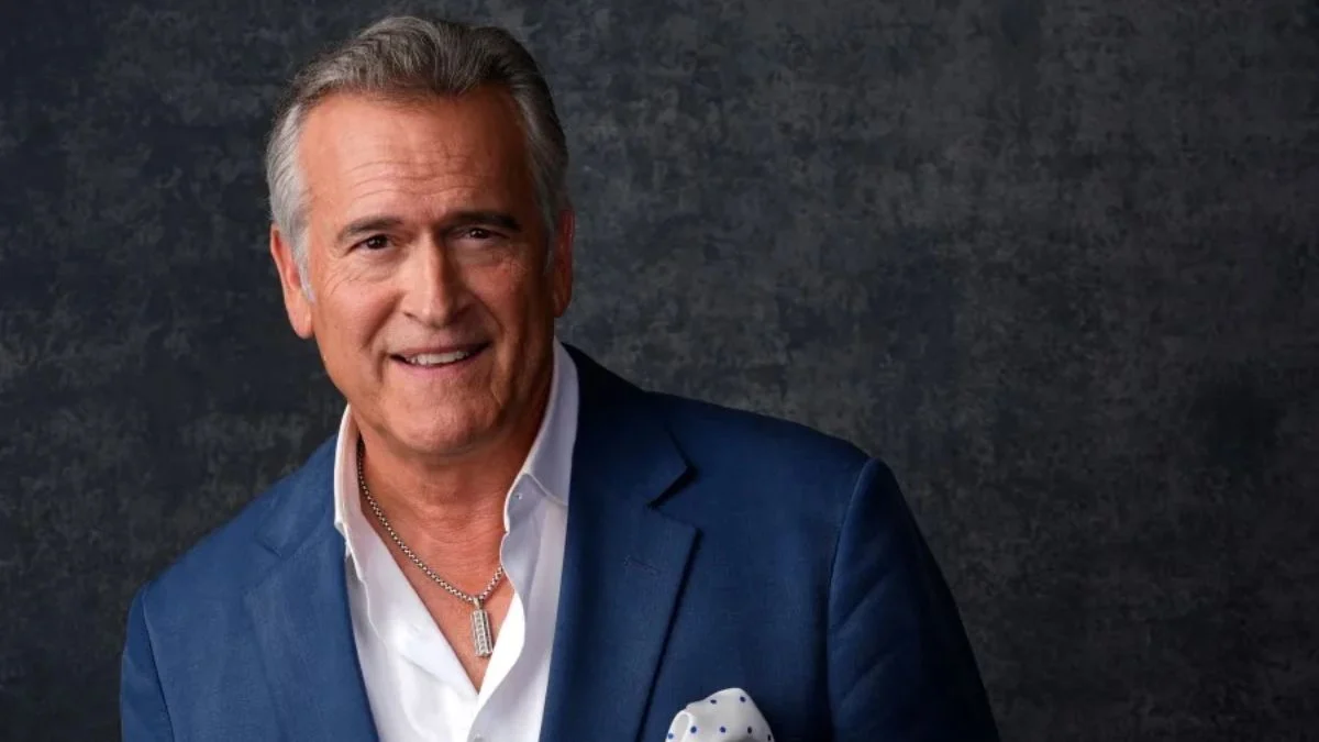 Actor Bruce Campbell wearing a blue suit jacket and white shirt smiling at a press event.
