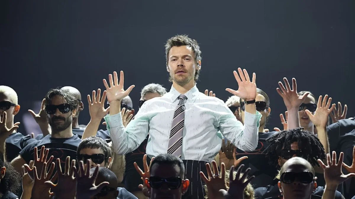 Harry Styles standing with hands raised surrounded by dancers in black shirts and sunglasses during a performance.