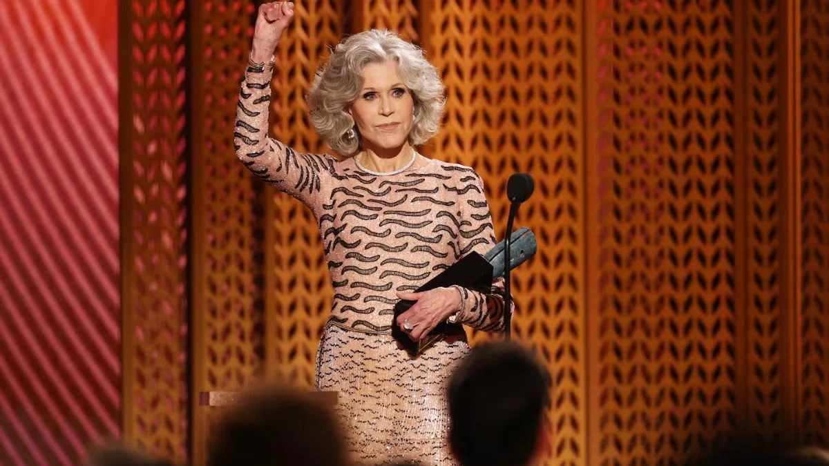 Jane Fonda standing at a podium with her fist raised in the air, holding an award during a televised ceremony.