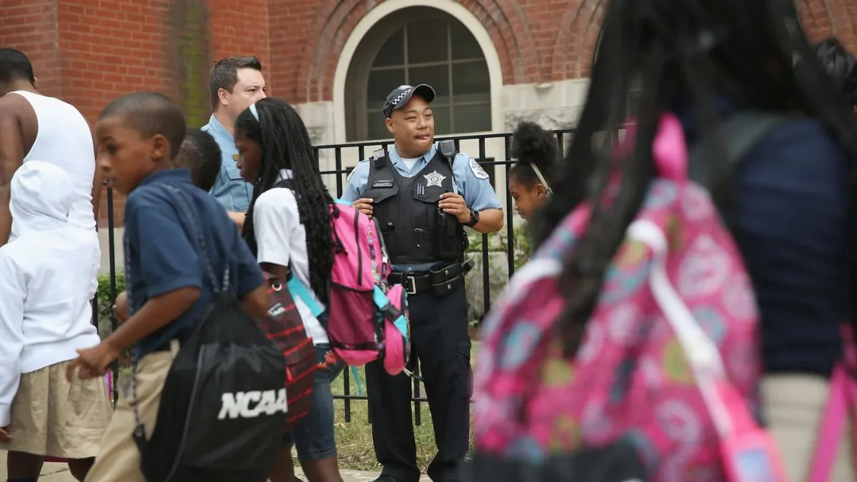 School resource officer standing in front of a brick building as students with backpacks walk by, illustrating the school-to-prison pipeline concept.