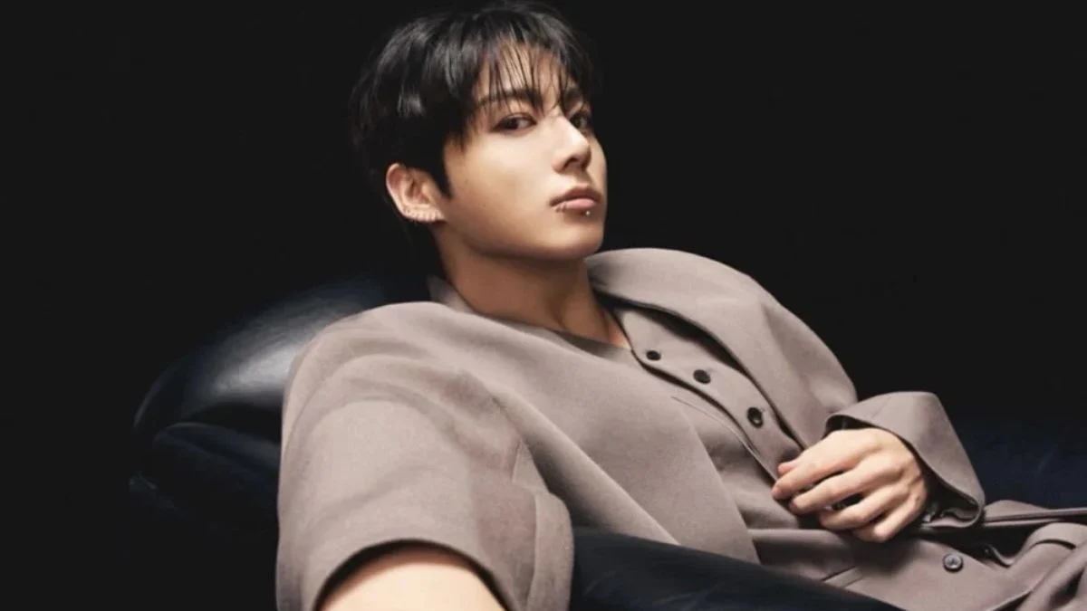 A high-fashion portrait of BTS member Jungkook reclining in a tan outfit against a dark background.