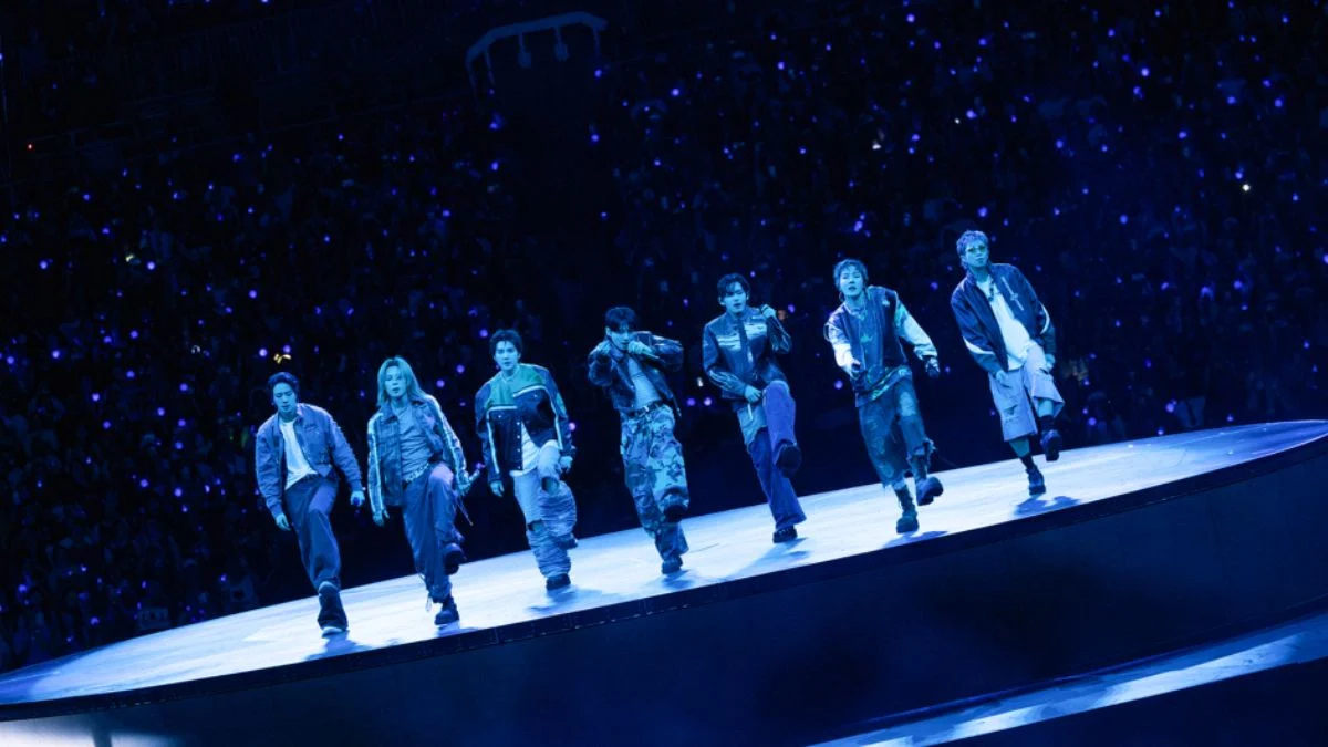 All seven members of K-pop group BTS walking across a large outdoor stadium stage under a blue lighting scheme, surrounded by an audience holding glowing blue lights.