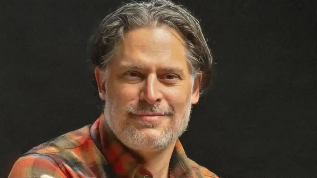 A close-up profile portrait of actor Joe Manganiello with graying hair and a trimmed beard, wearing a plaid shirt against a dark background.