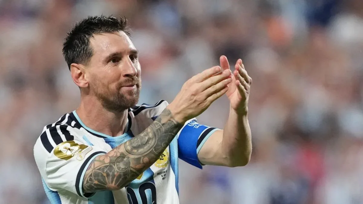 Lionel Messi clapping his hands and smiling on the pitch while wearing Argentina's white and light-blue striped jersey with a captain's armband during a World Cup match.
