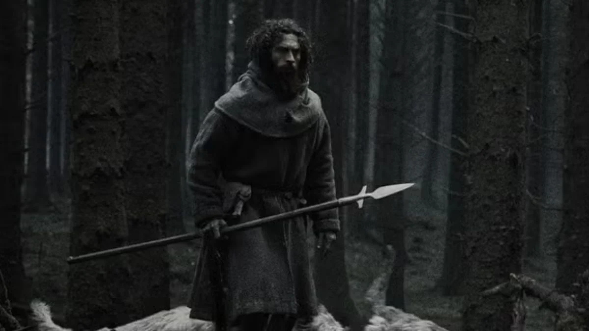 Actor Aaron Taylor-Johnson with long dark hair and a beard, dressed in a rugged 13th-century medieval hooded tunic, holding a spear while standing in a dark, foggy forest.