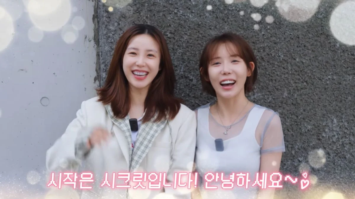SECRET members Jun Hyoseong and Jung Hana smiling together in front of a grey textured wall in their first official reunion video presentation on YouTube.