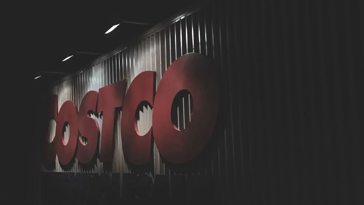 An illuminated outdoor storefront sign featuring the large red letters of the Costco Wholesale corporate logo on a corrugated metal wall under dim night lighting.