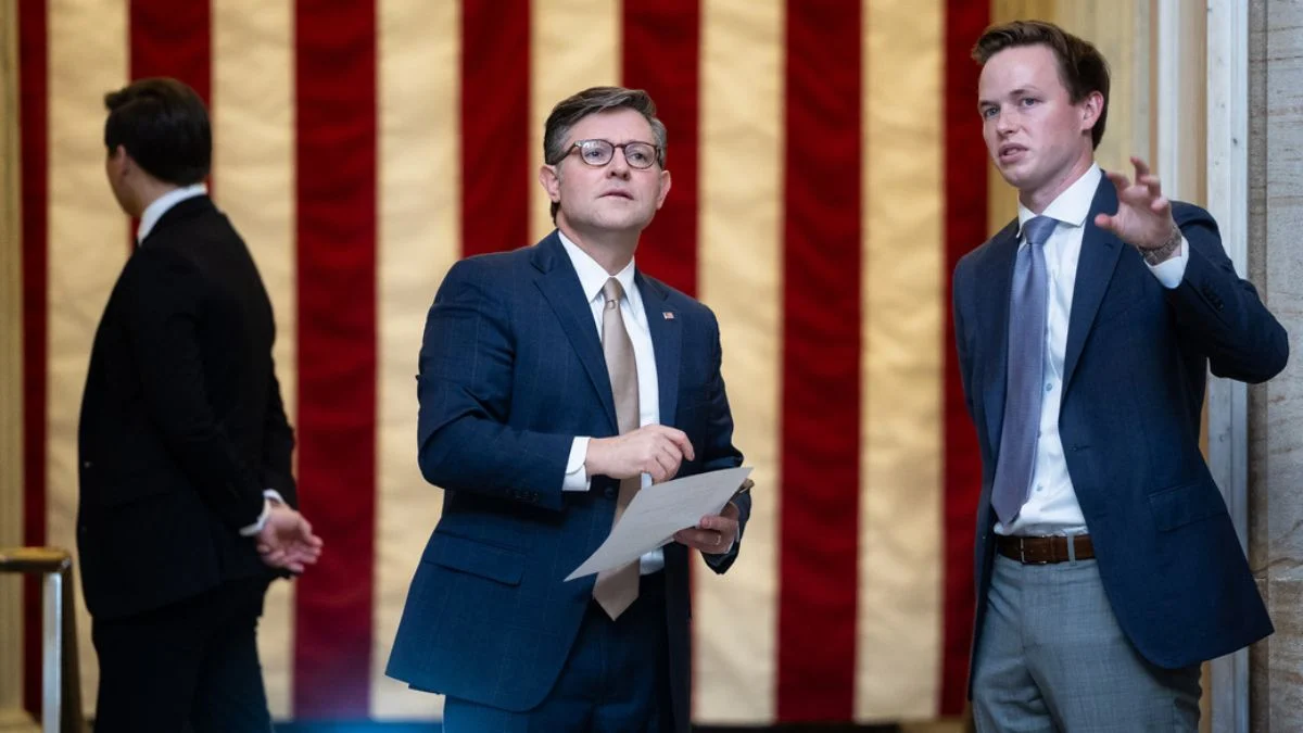 Speaker Mike Johnson holding papers and speaking with an aide in front of a large American flag backdrop in the US Capitol.